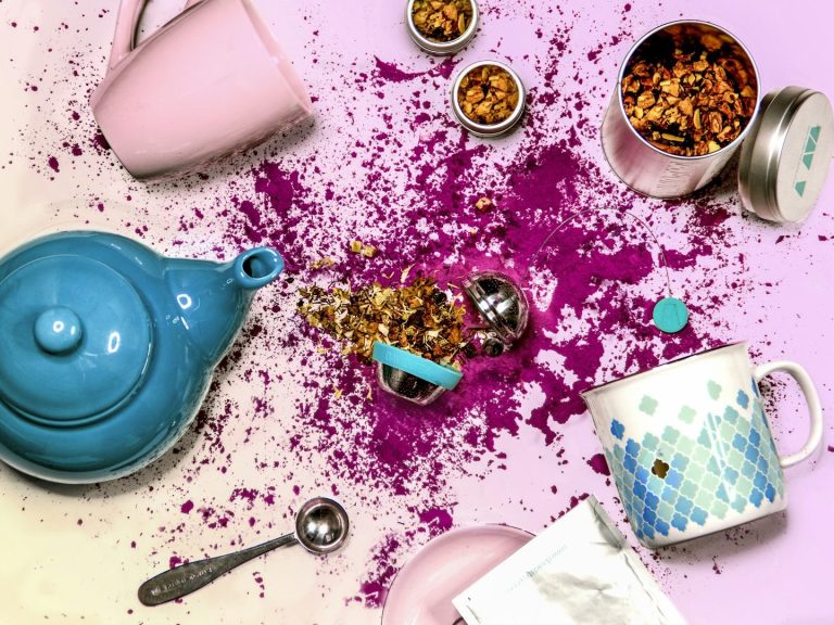 Tetera azul, tazas, especias y pétalos de flores sobre una superficie blanca con manchas de color.